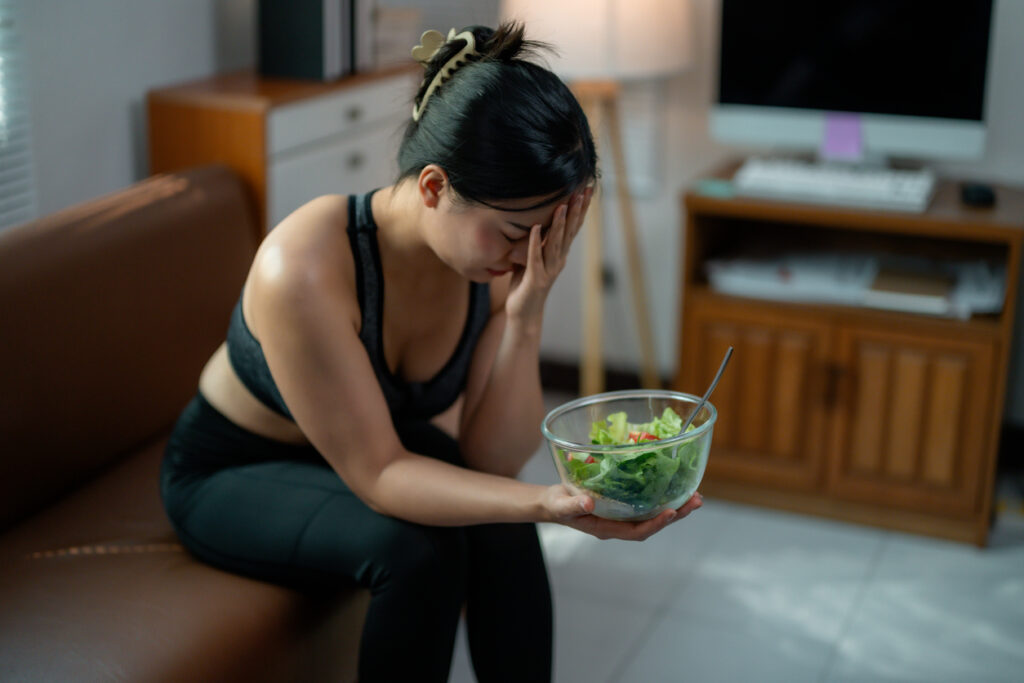 Beyond Calories: Hidden Reasons for Weight Gain, Young Asian woman feeling bored and tired while eating a healthy salad at home, sitting on the sofa in her living room, she's following a strict diet