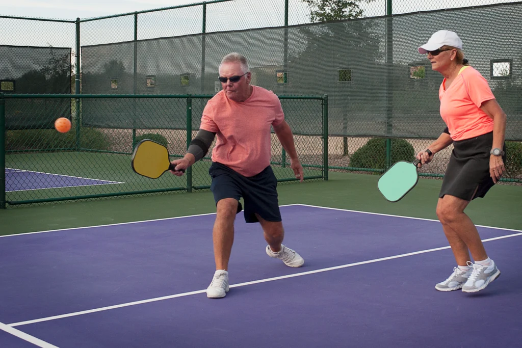 Active senior man hitting an orange pickleball on a purple court with a senior woman ready to play.