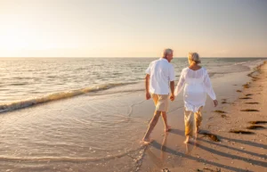 Elderly couple holding hands walking on a sandy beach at sunset.