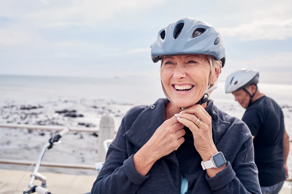Older woman fastening her bicycle helmet while smiling near the waterfront, representing strength, mobility, and healthy aging through an active lifestyle.