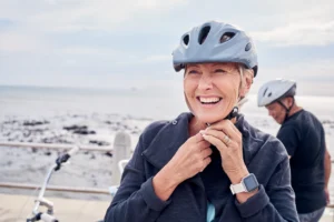 Older woman fastening her bicycle helmet while smiling near the waterfront, representing strength, mobility, and healthy aging through an active lifestyle.
