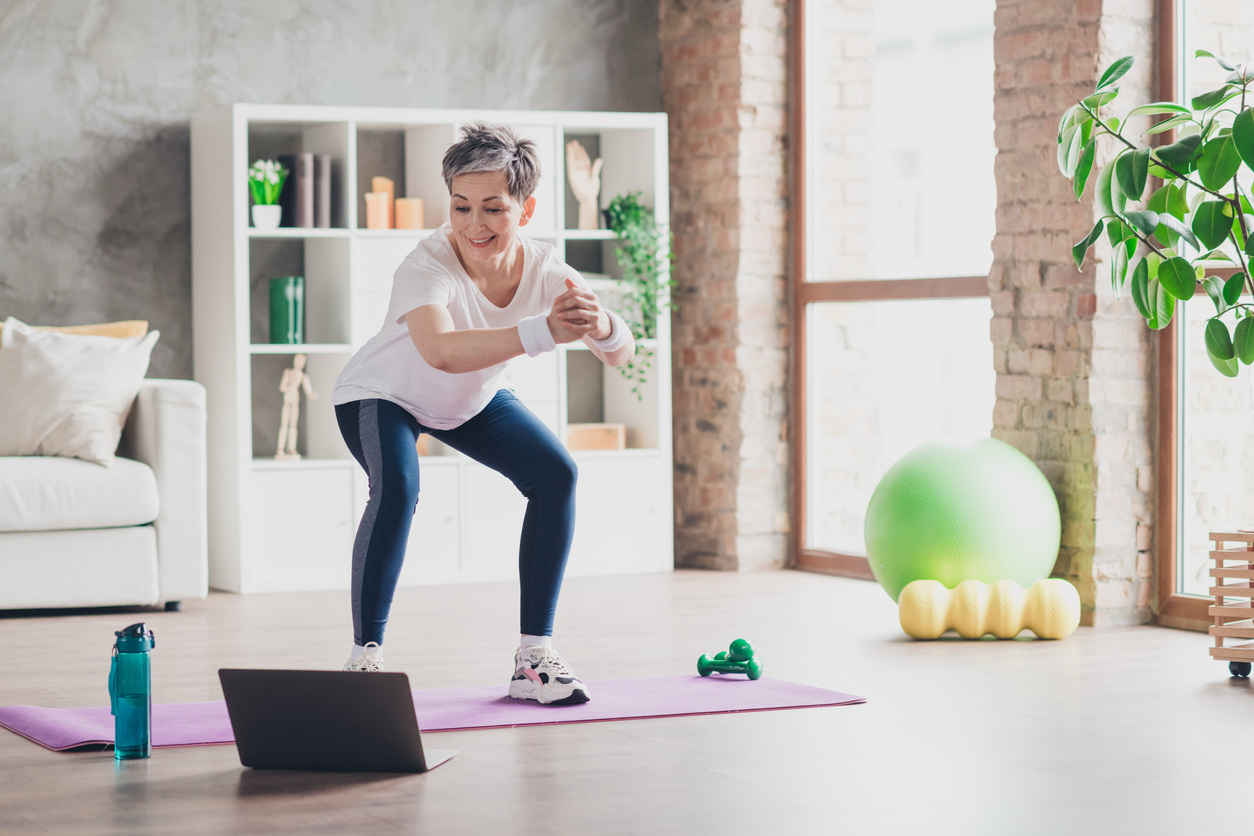 Woman following an online workout while performing bodyweight squats at home to build strength for healthy aging.