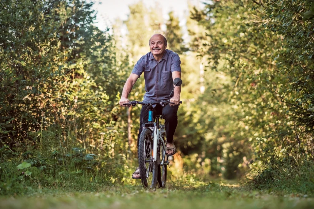 Older man riding a bicycle along a wooded trail, illustrating the strength, mobility, and independence that support healthy aging.