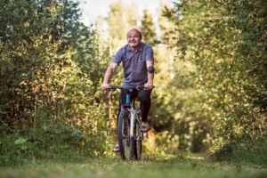 Older man riding a bicycle along a wooded trail, illustrating the strength, mobility, and independence that support healthy aging.